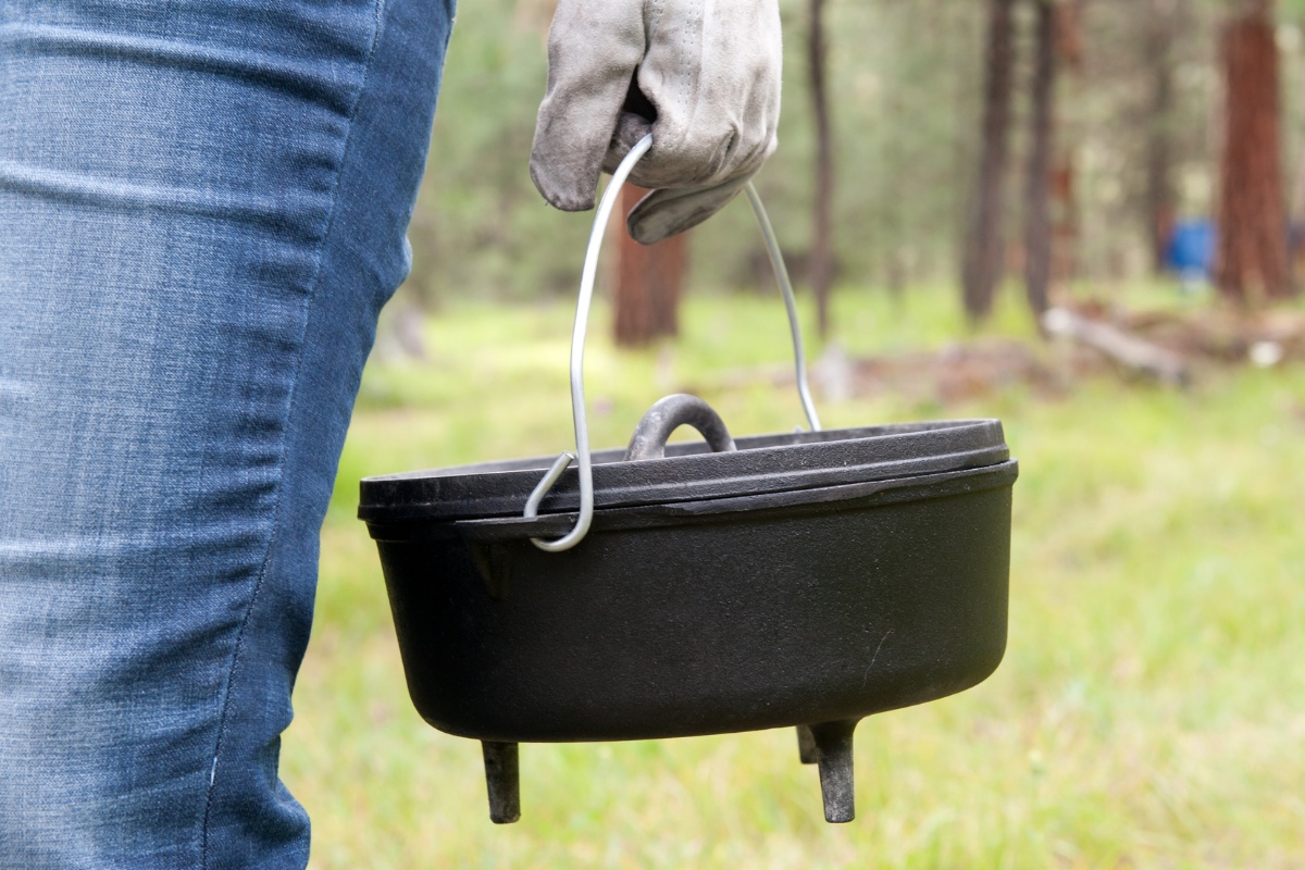 Nahaufnahme eines Dutch Ovens, der von einer Person mit Handschuh getragen wird, im Hintergrund Bäume.