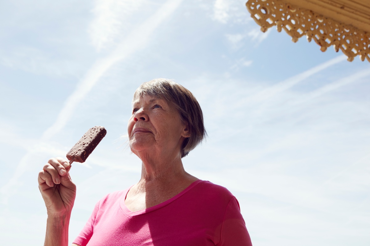 Eine ältere Frau genießt ein Schokoladeneis am Stiel unter blauem Himmel.