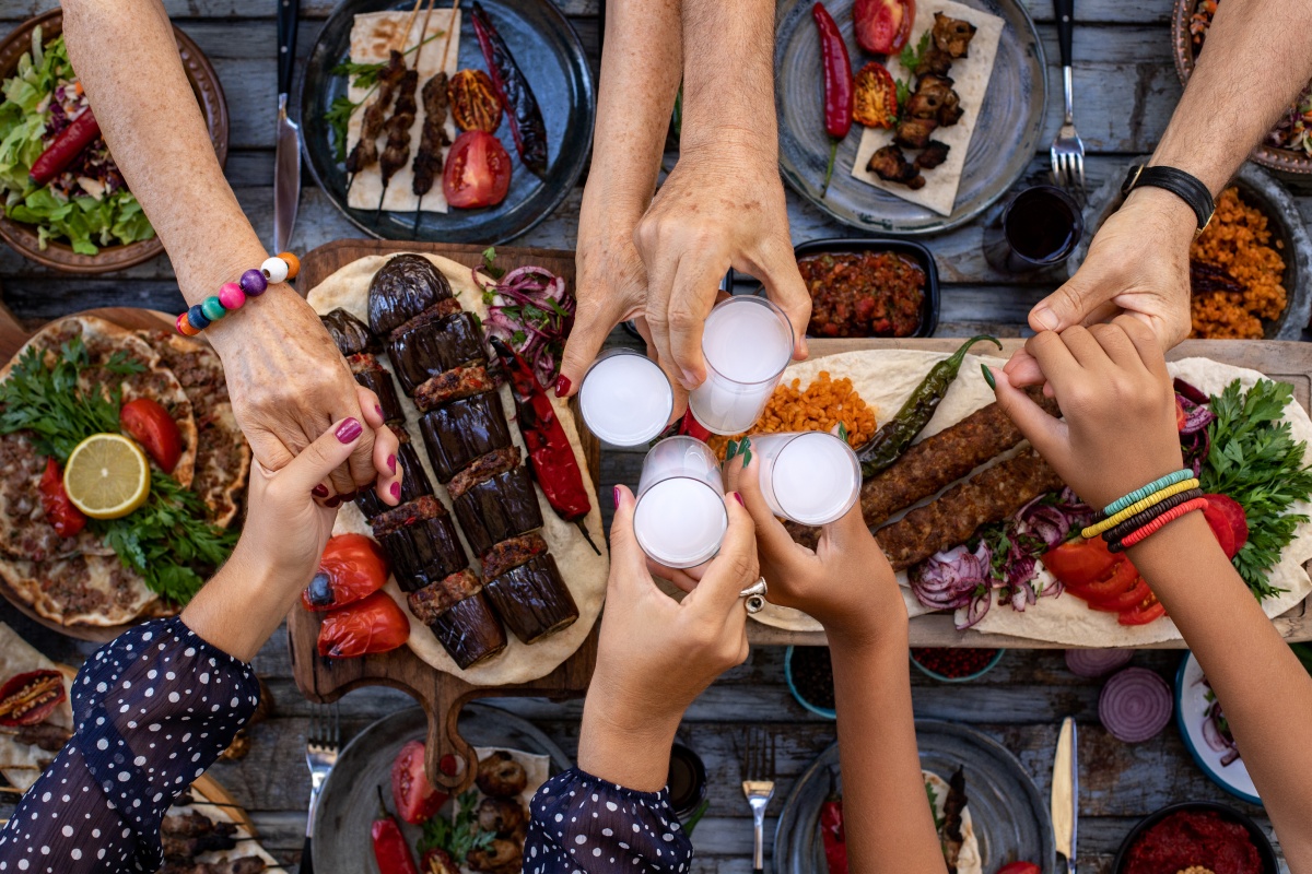 Personen halten sich bei den Händen und stoßen mit Ouzo über einem Tisch mit griechischen Spezialitären an.