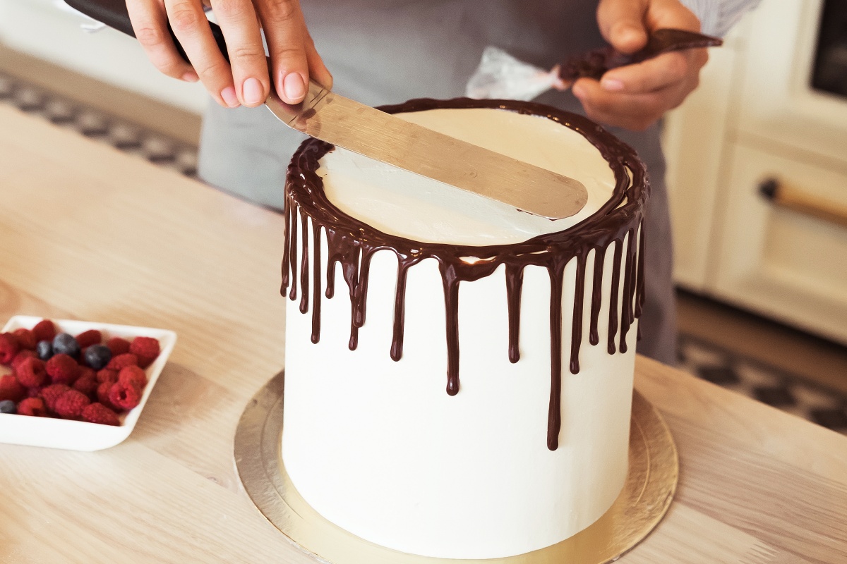 Konditor glättet weiße Torte mit am Tortenrand herabtropfender Schokoladenglasur.