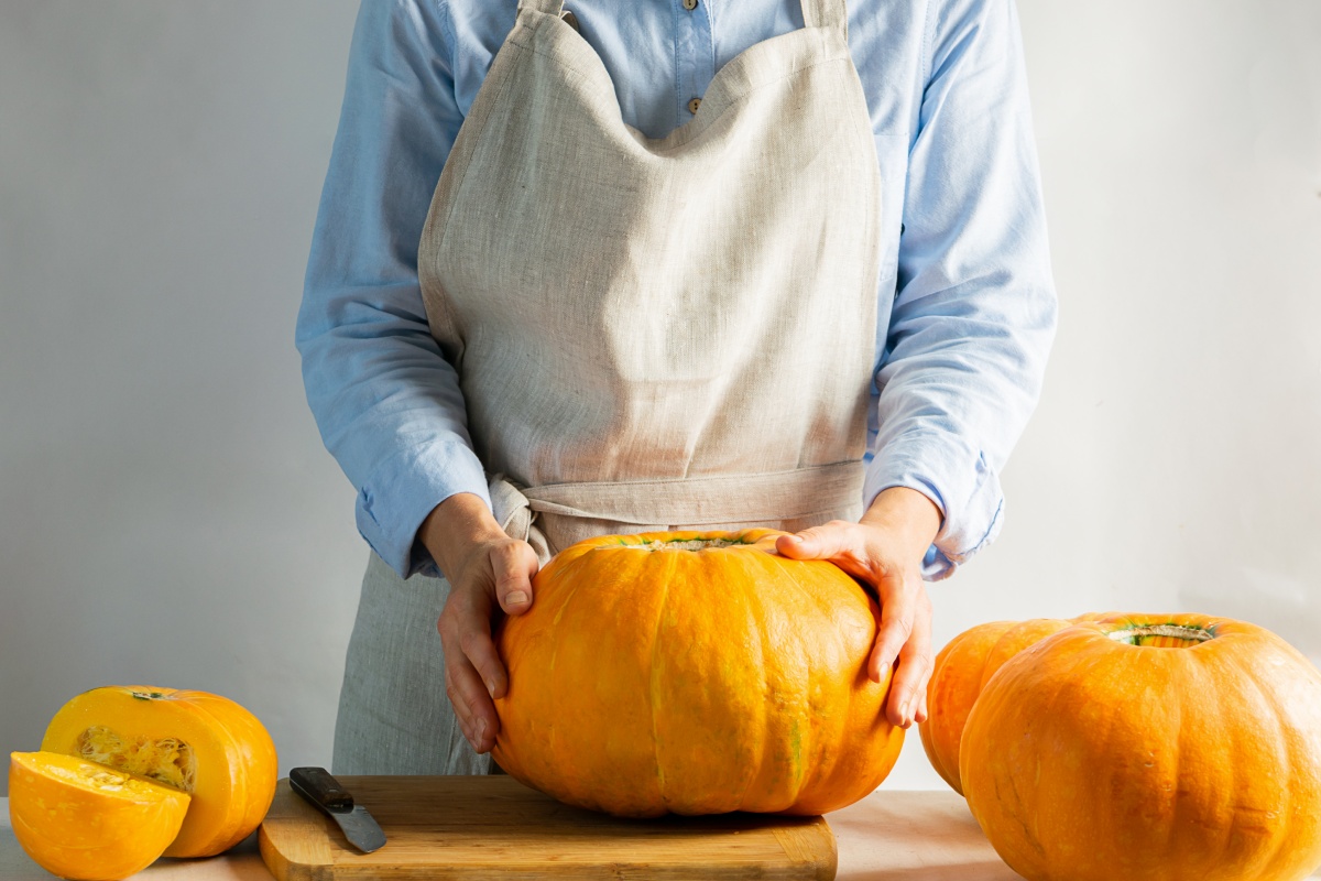 Person in Schürze hält einen großen, runden Kürbis auf einem Schneidebrett. Weitere Kürbisse und Messer daneben.