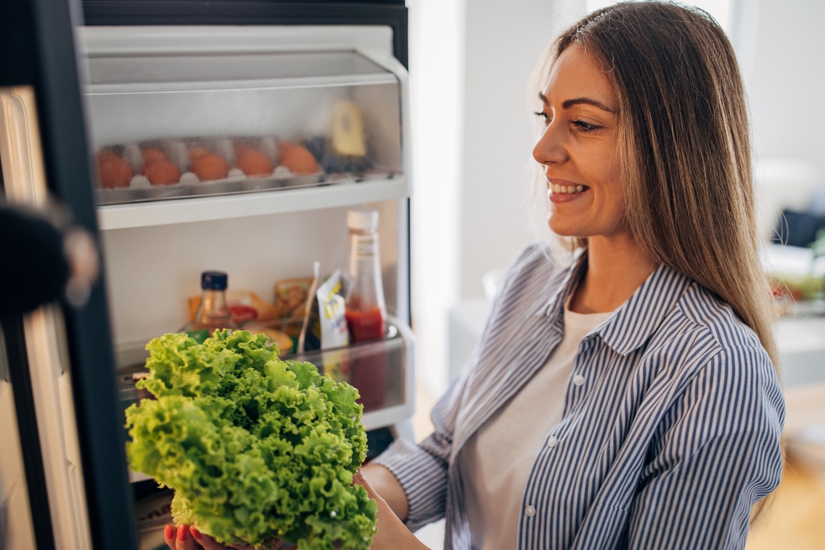 Lächelnde Frau steht vor einem geöffneten Kühlschrank und hält frischen grünen Salat in der Hand.