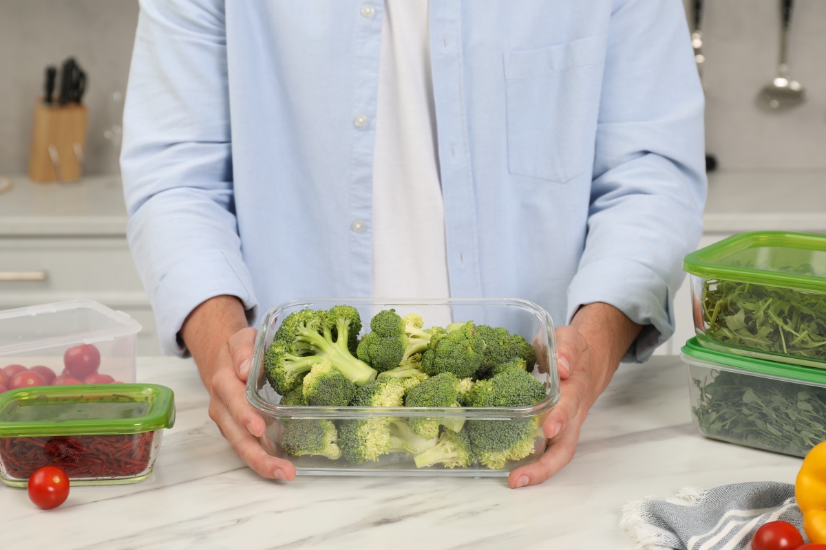 Person hält eine Frischhaltebox mit frischem Brokkoli, umgeben von weiteren Gemüsebehältern in der Küche.