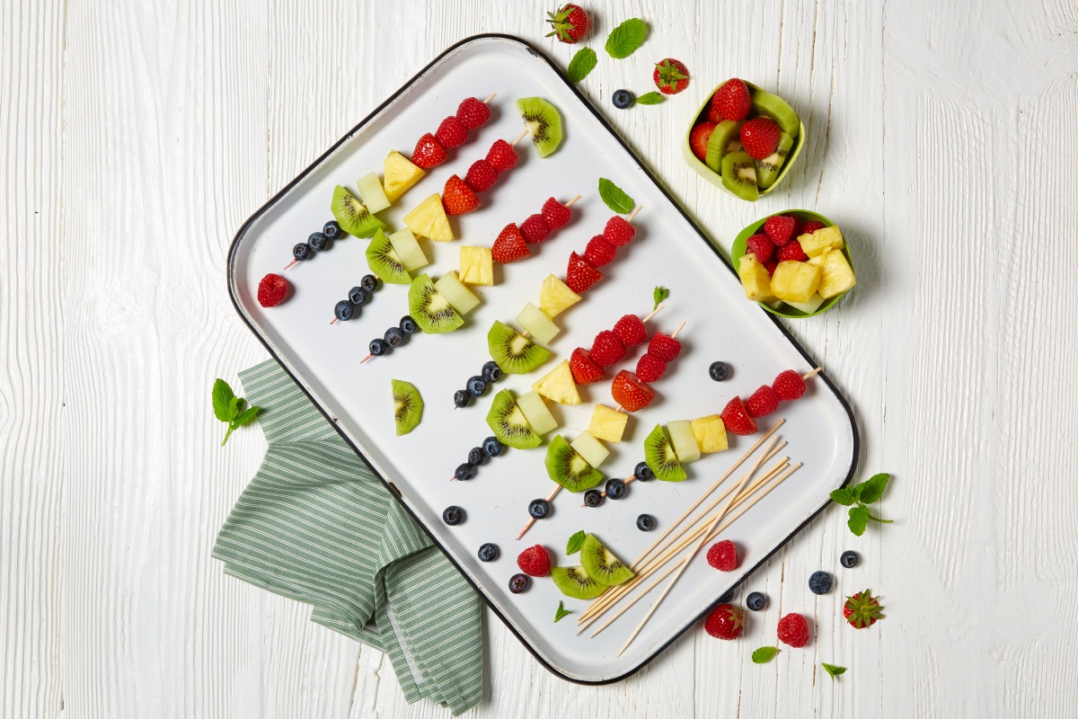 Bunte Obst-Spieße aus Kiwi, Ananas und Himbeeren liegen auf einem weißen Tablett.