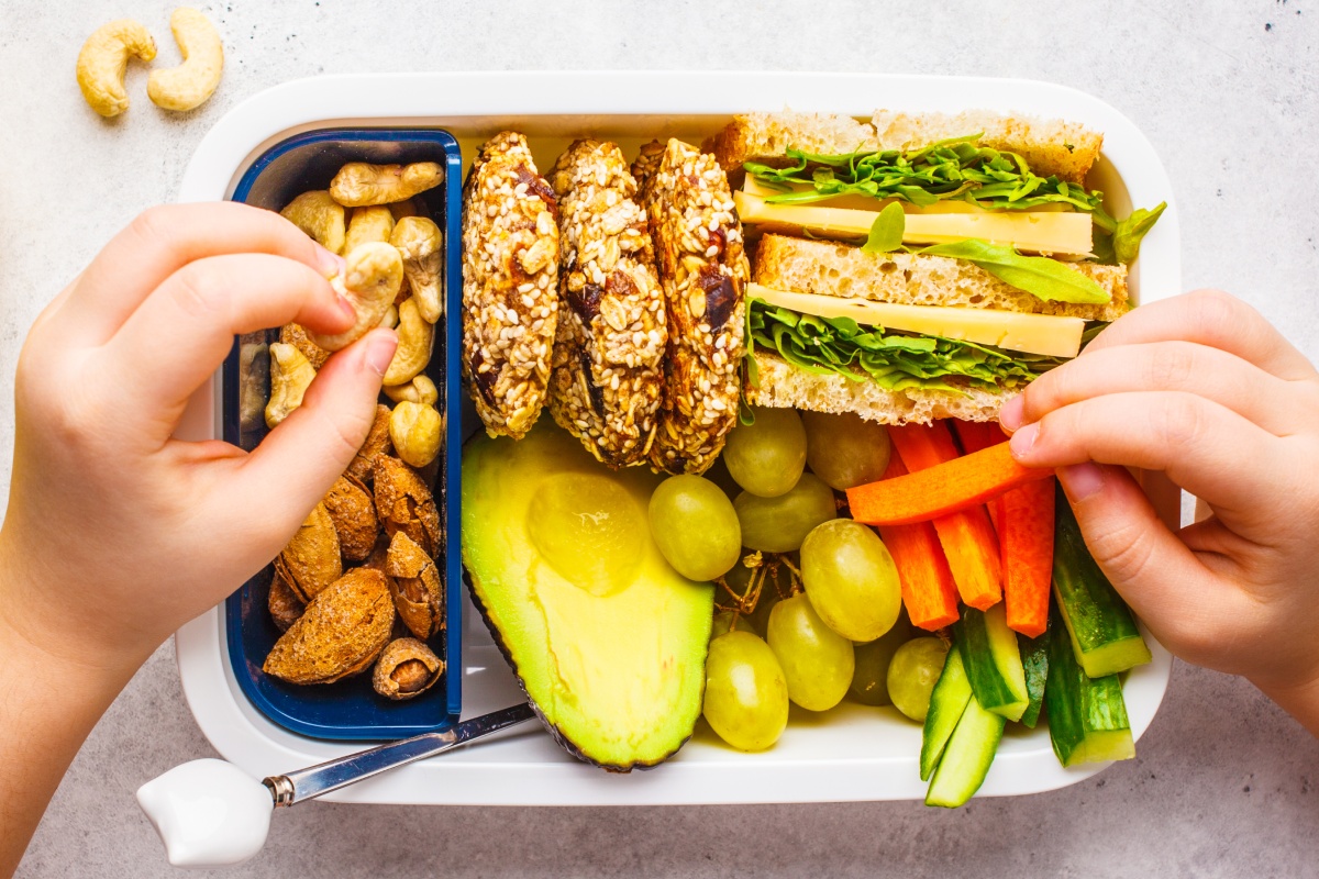 Ein Kind nimmt sich Nüsse und Möhrensticks aus einer bunt gefüllten Brotdose, in der sich noch Obst, Sandwichse, Gurke und Snacks befinden.