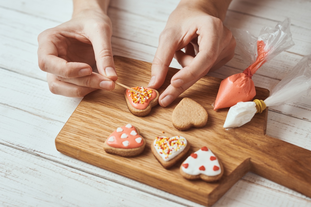 Jemand verziert gebackene herzförmige Plätzchen mit rosa Zuckerguss und Streuseln auf Holzbrett.