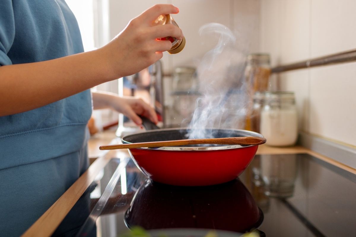 Person würzt ein Gericht in einer dampfenden roten Pfanne auf dem Herd. Holzlöffel liegt auf dem Rand.