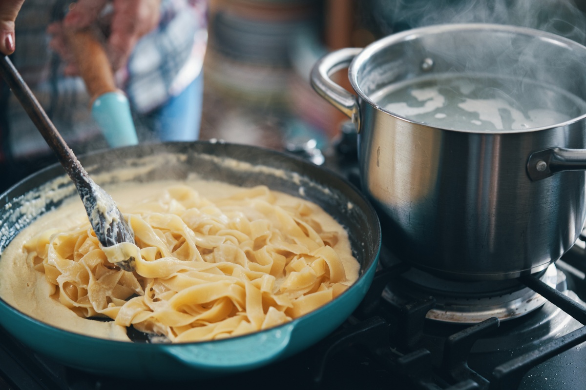 Tagliatelle in cremiger Sauce in einer Pfanne, daneben ein dampfender Kochtopf auf dem Herd.