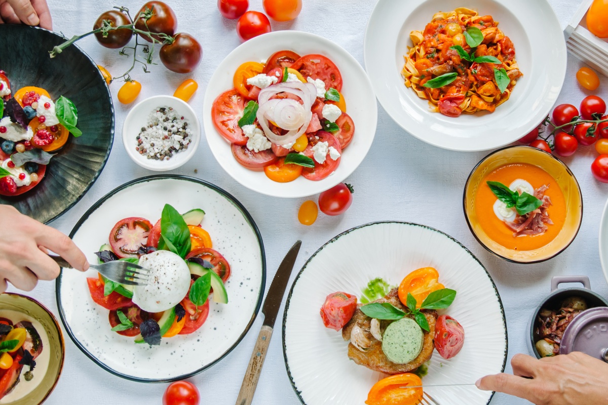 Draufsicht eines Tisches mit weißer Tischdecke und vielen Tellern und Schalen, die mit aufgeschnittenen Tomaten und verschiedenen Tomatenspeisen gefüllt sind.