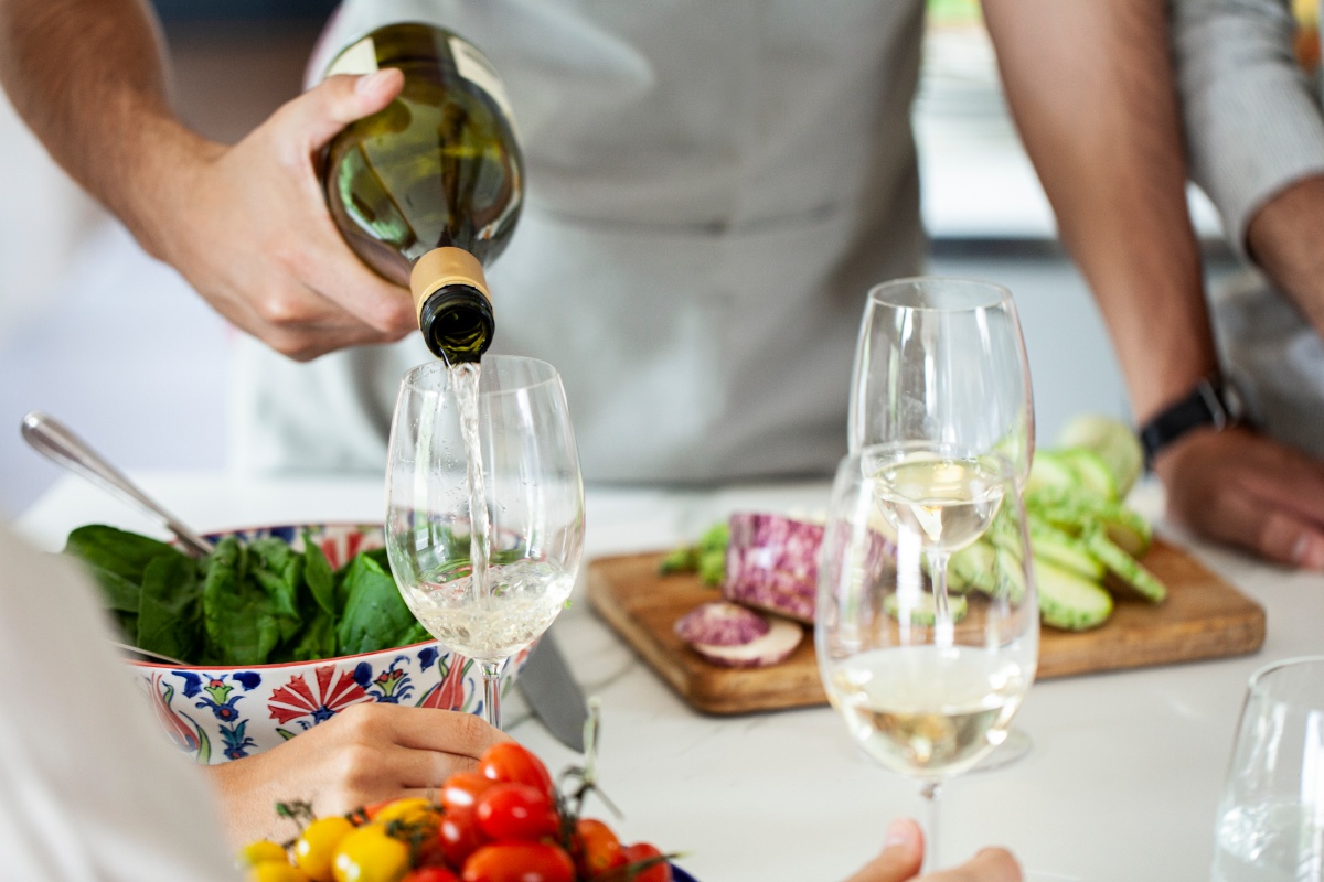 Person gießt Weißwein in ein Glas, während eine Schüssel Salat und geschnittenes Gemüse auf einem Tisch stehen.