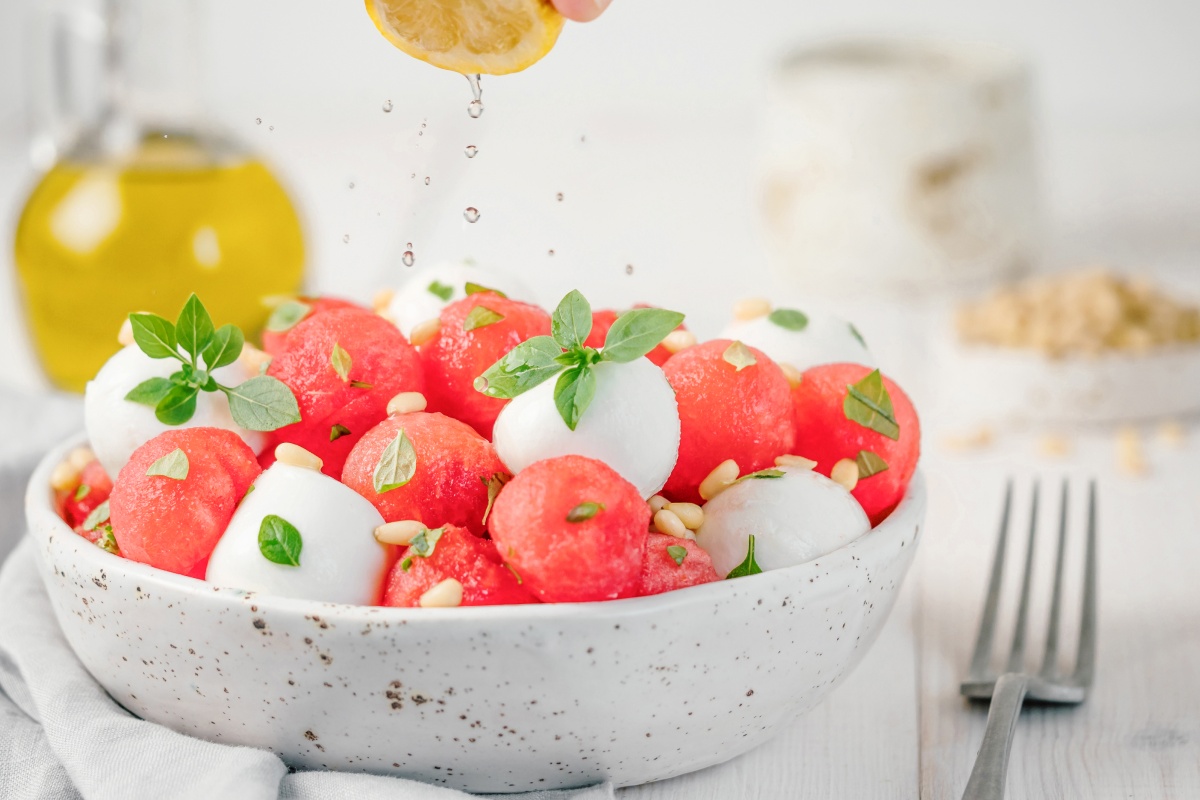Ein Melonen Mozzarella Salat, garniert mit Pinienkernen und Basilikum, wird mit frisch gepresster Zitrone übergossen