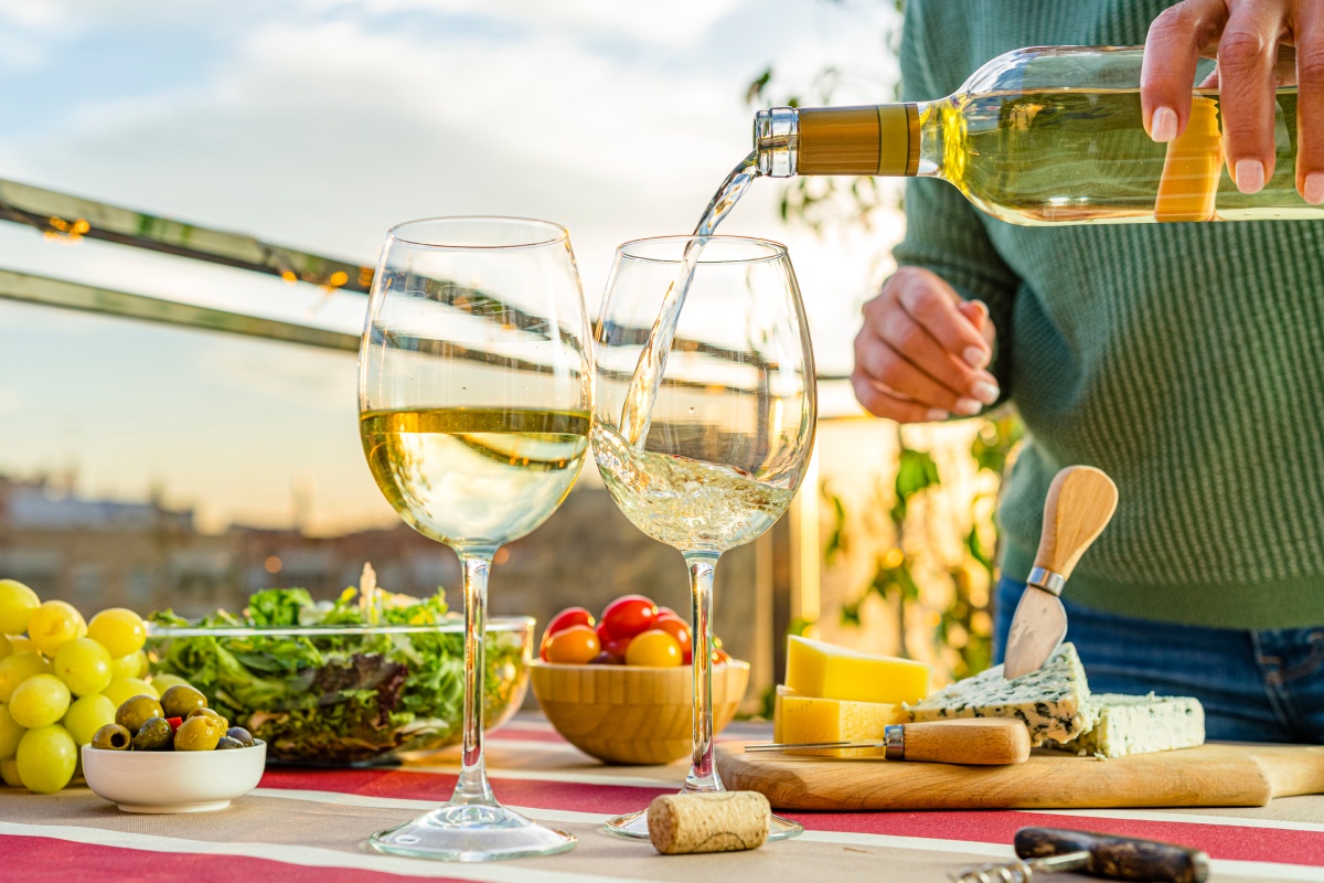 Person schenkt Weißwein in zwei Gläser auf einem Tisch mit Käse, Trauben und Salat bei Sonnenuntergang ein.