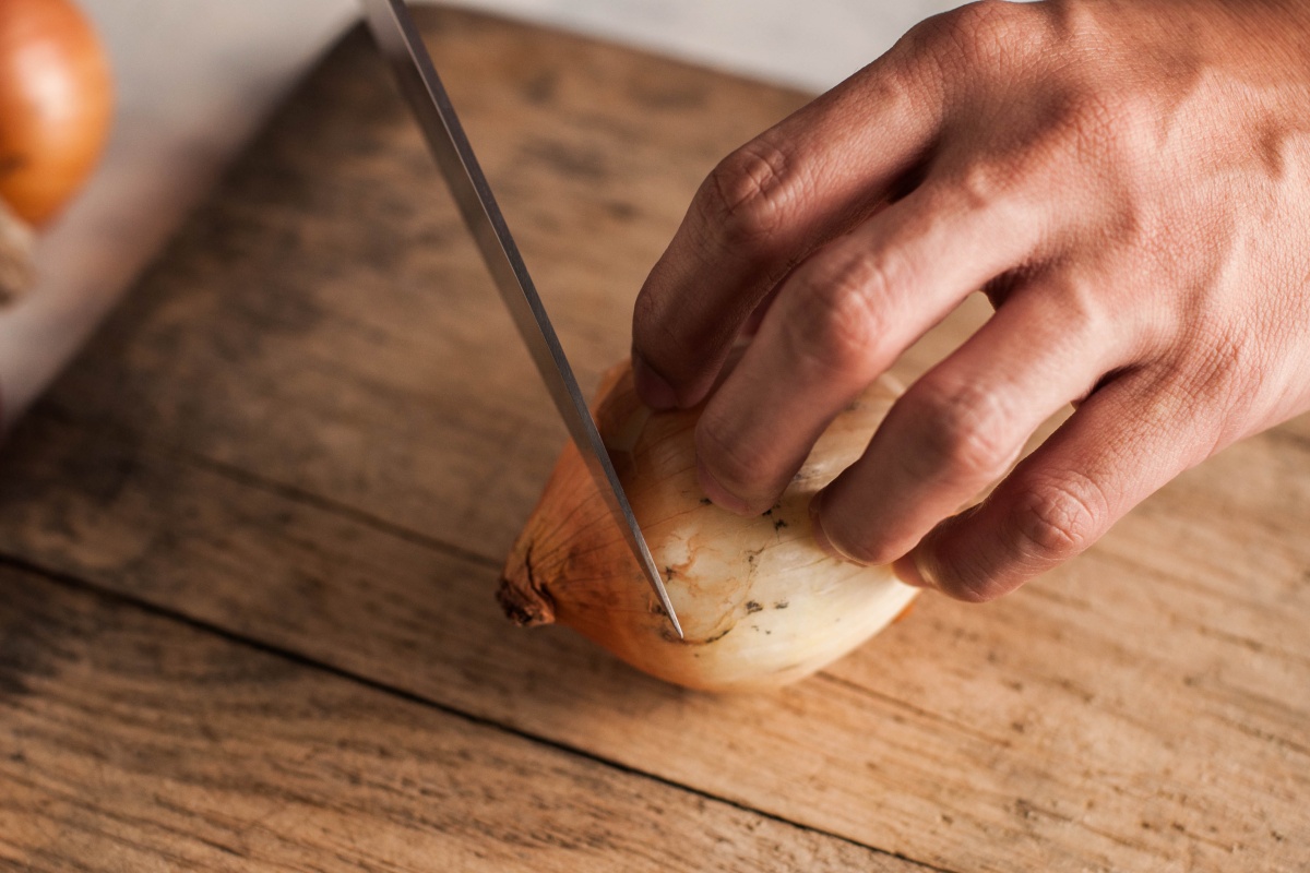 Person schneidet eine ungeschälte Zwiebel auf einem rustikalen Holzbrett mit einem scharfen Messer.