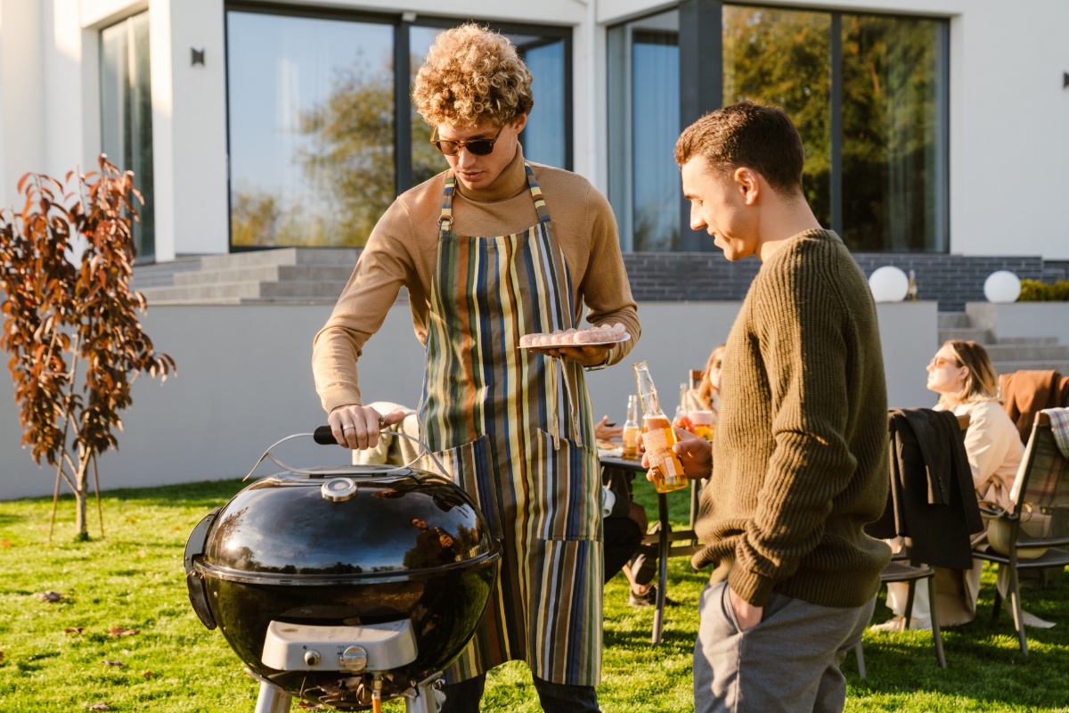 Zwei Männer stehen draußen am Grill, einer mit Sonnenbrille, Schürze und einem Teller mit ungegrillten Würstchen in der Hand, daneben einer eine Flasche Limonade haltend. Im Hintergrund gedeckter Tisch mit Menschen und ein Haus.