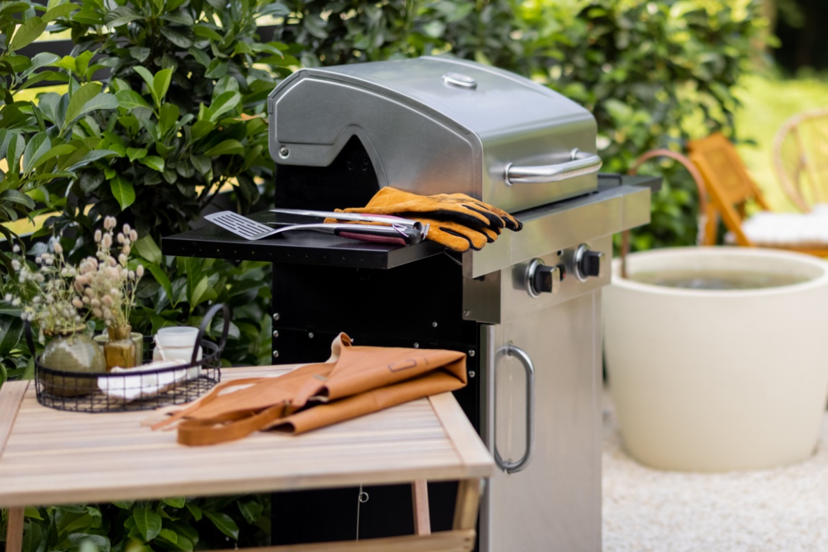 Kleiner Holztisch mit gefalteter Schürze darauf, daneben Gasgrill mit Grillhanzschuhen und Grillbesteck, dahinter Heckensträucher.