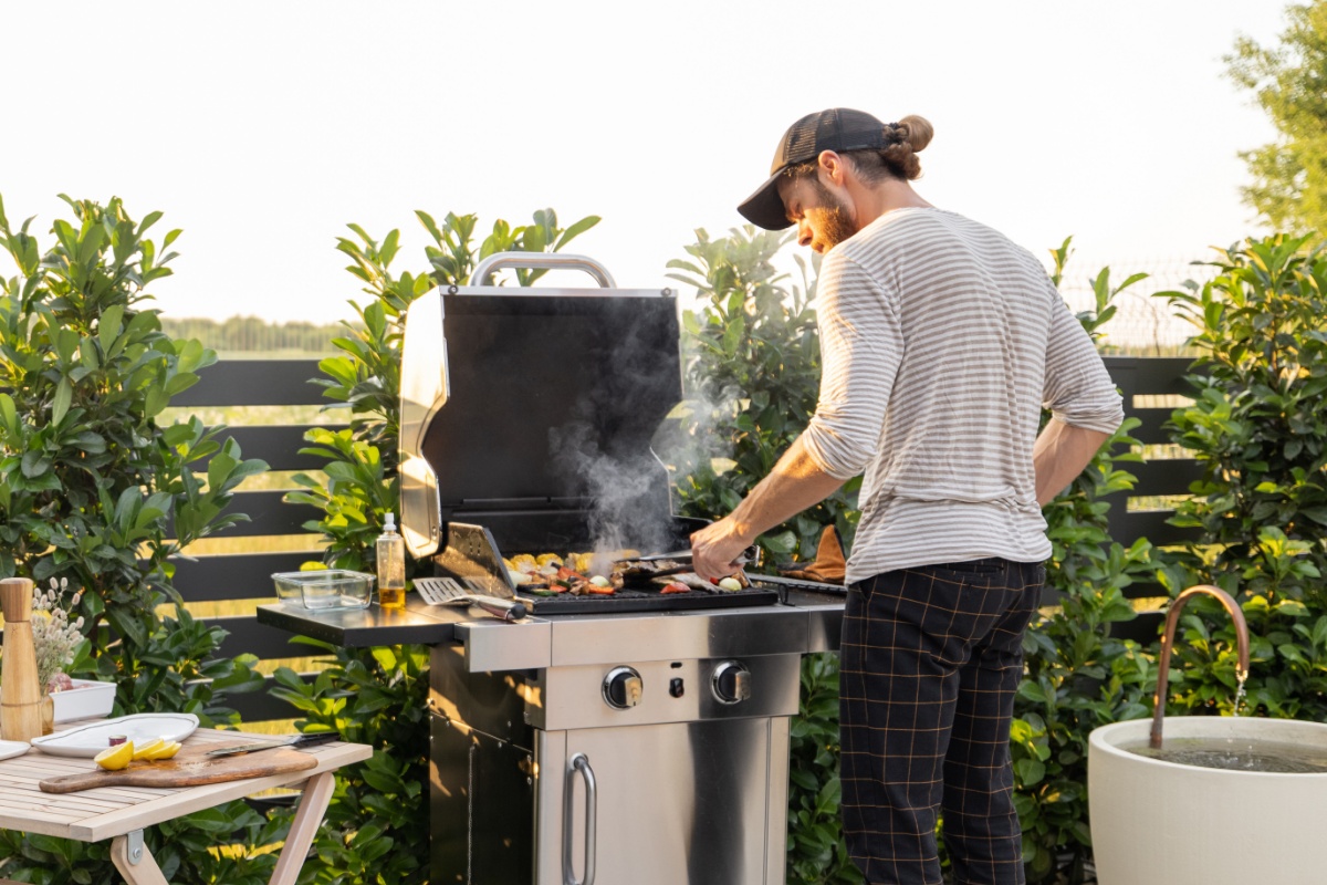 Mann in gestreiftem Shirt, karierter Hose und Schildmütze steht mit Grillzange am mit Gemüse belegten, dampfenden Gasgrill. Auf der Ablage des Grills stehen Ölflasche, leere Aufbewahrungsbox und Pfannenwender. Links davon ein kleiner Holz-Gartentisch mit Schneidebrett und Messer, dahinter Gartenzaun mit Heckensträuchern, dahinter Feld und Bäume.