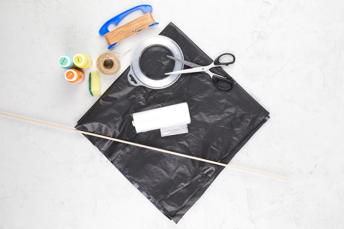 Materialien zum Drachen basteln. Schwarze Müllsäcke, Schere, Klebeband, Schnur, Holzstäbe, Farben und Drachenleine auf hellem Hintergrund.