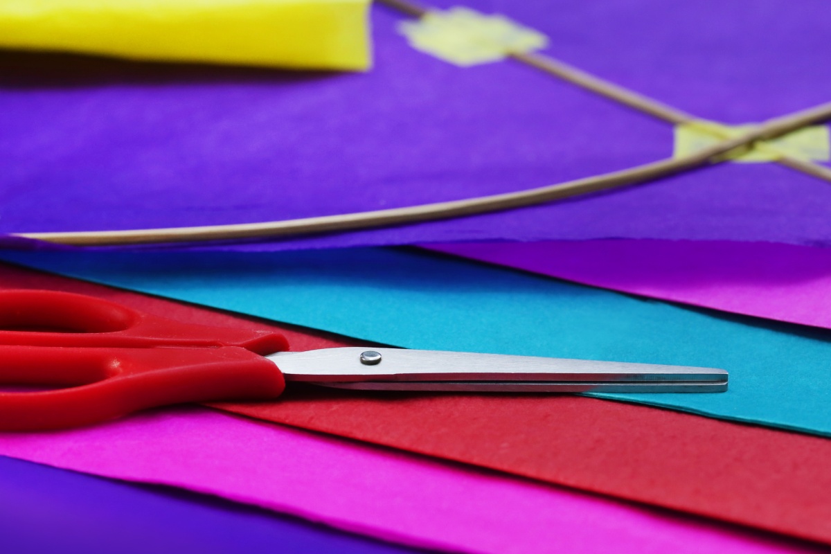 Rote Schere liegt auf buntem Papier neben einem teilmontierten Drachen aus Holzstäben, bereit zum Basteln.