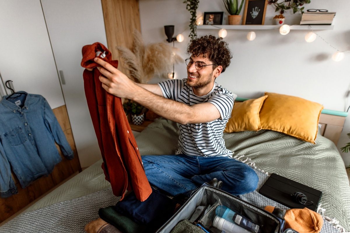 Mann sitzt auf einem Bett beim Kofferpacken. Er hält ein rotes Hemd hoch.
