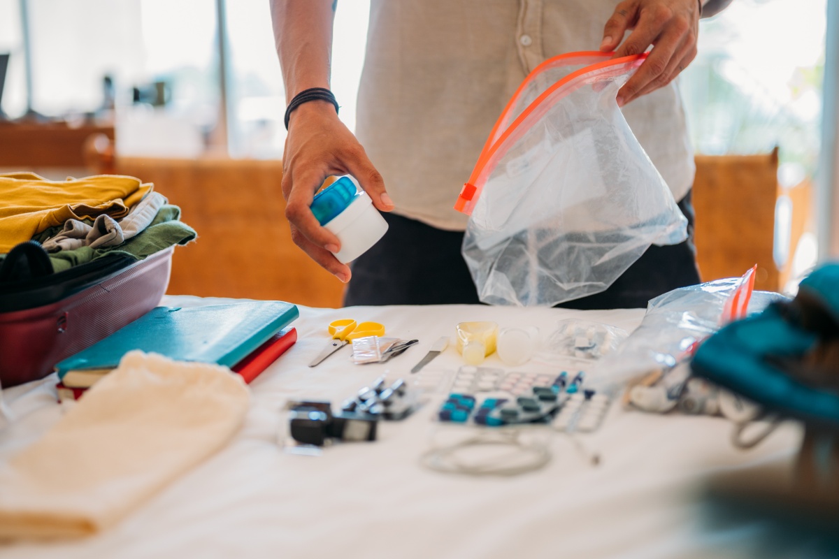 Person packt kleine Gegenstände in einen durchsichtigen Beutel, daneben liegen Kleidung und Bücher.