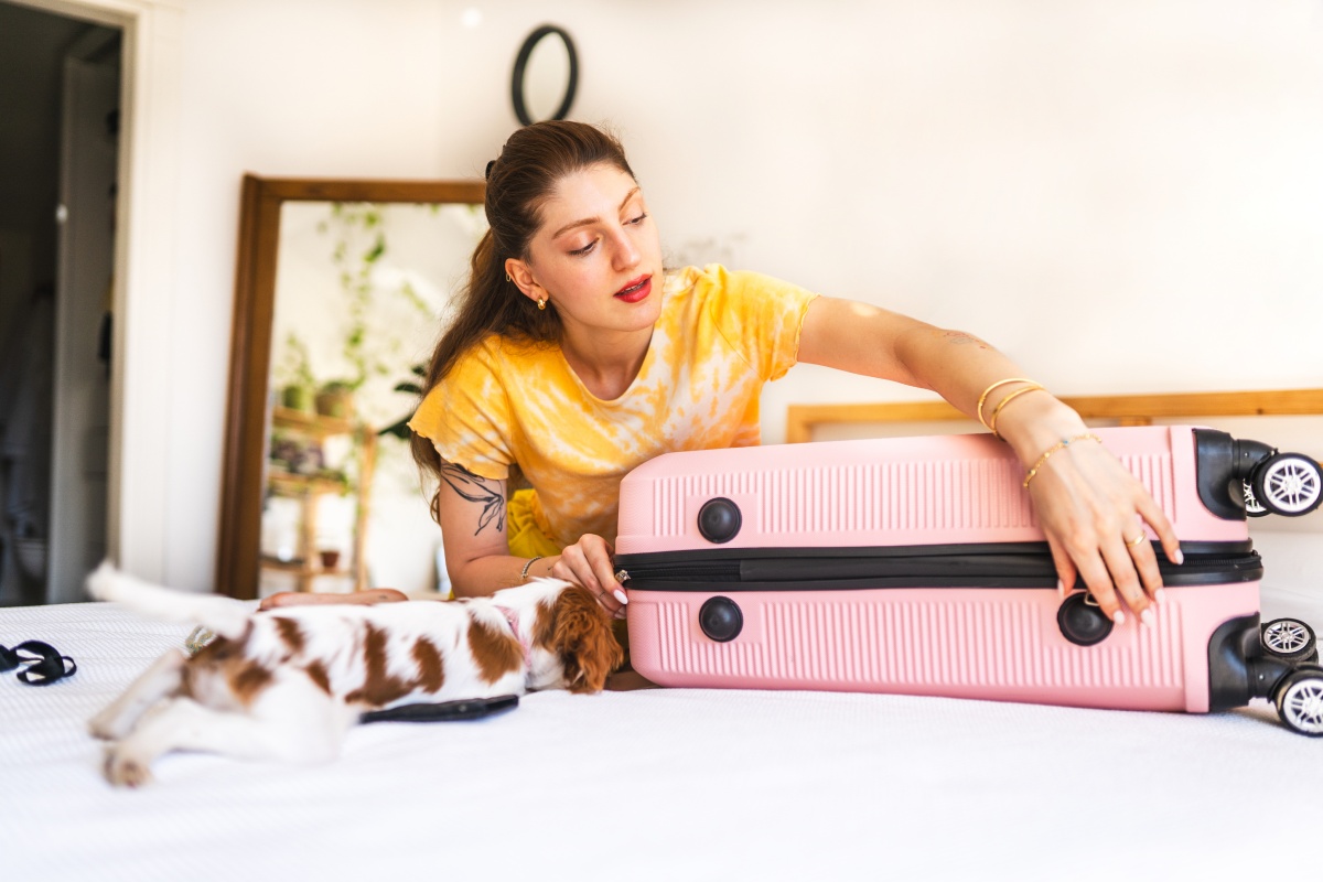 Frau schließt ihren gepackten Koffer auf einem Bett, während ein kleiner Hund daneben liegt.