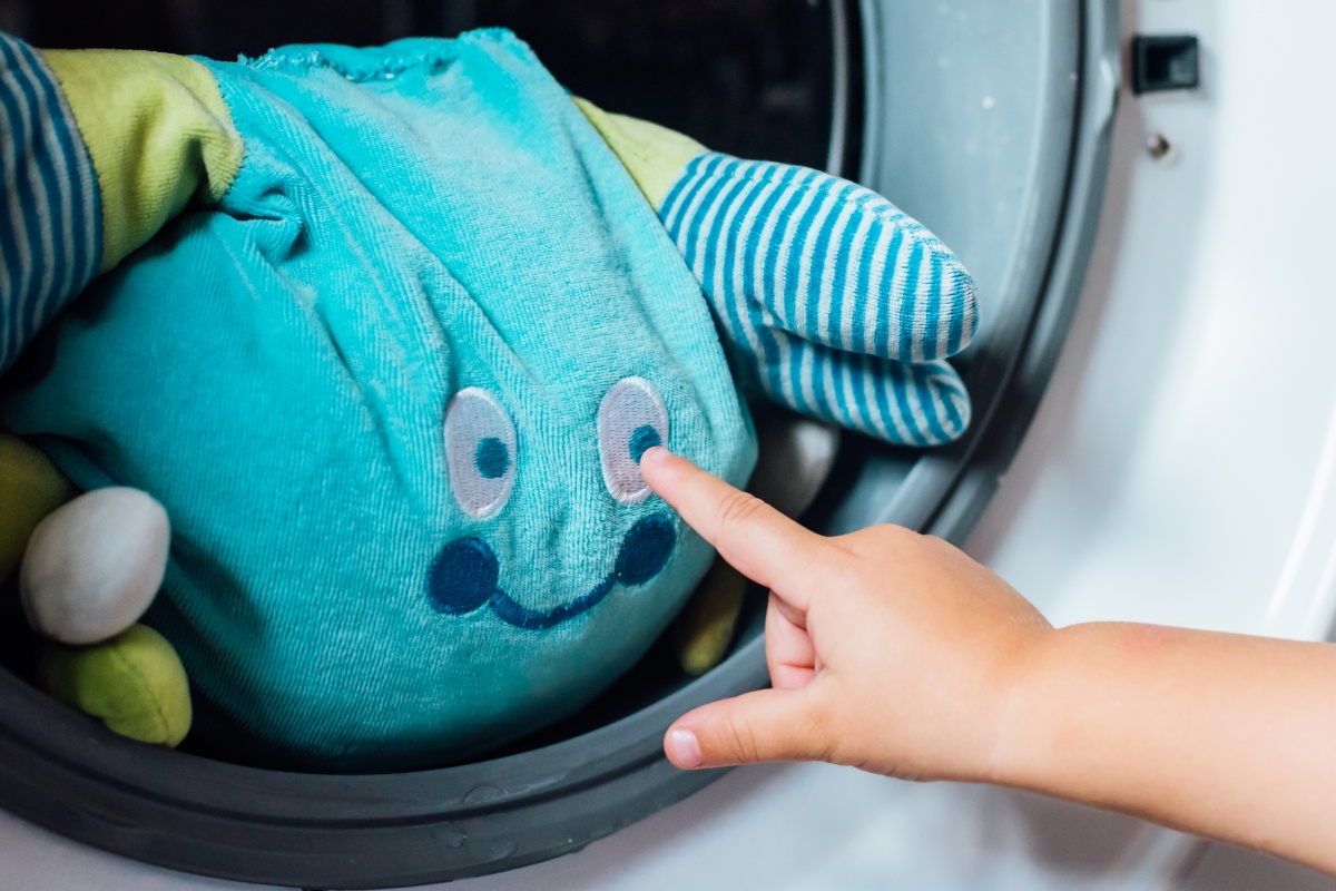 Ein Kuscheltier schaut aus der geöffneten Waschmaschine, während ein Kind ihm einen Stubser gibt.