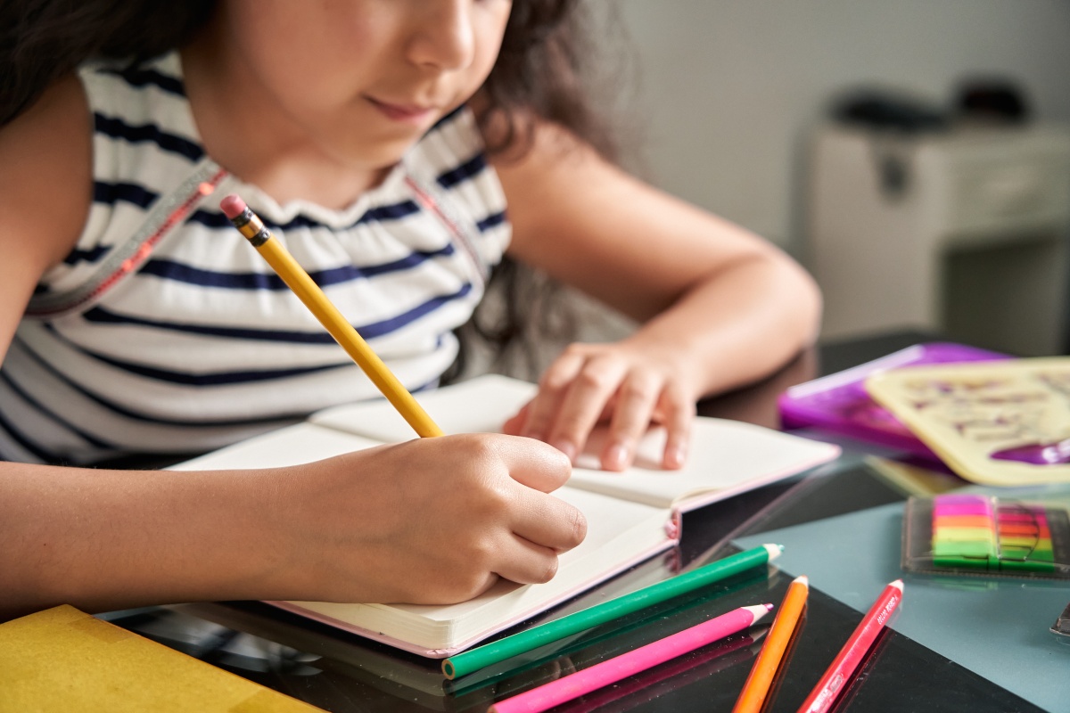 Mädchen in gestreiftem Shirt schreibt in ein Notizbuch, auf dem Tisch liegen Buntstifte und weitere Schreibutensilien.