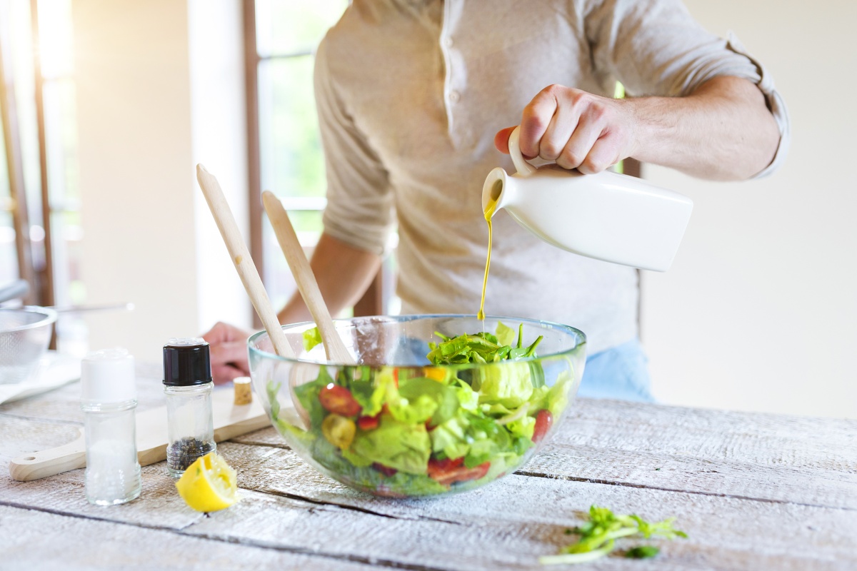 Person gießt Olivenöl in eine Schüssel mit frischem Salat auf einem Holztisch, Gewürze und Zitrone daneben.