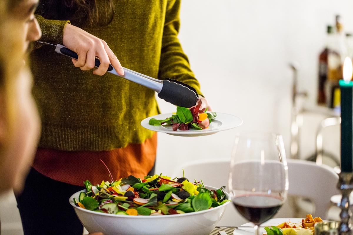 Person serviert frischen Salat auf einen Teller bei einem Abendessen, daneben steht ein Glas Rotwein.
