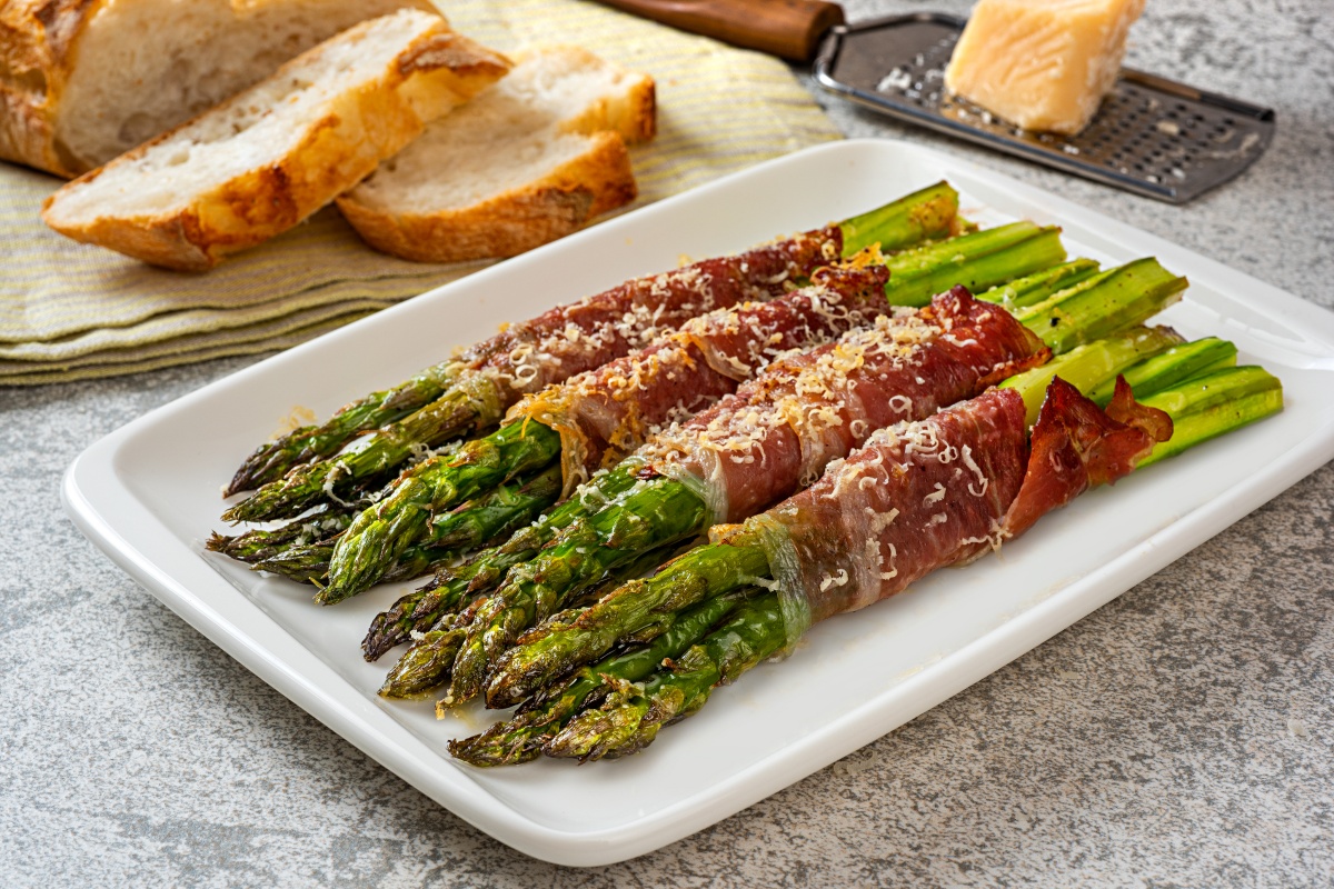 In Speck gewickelter grüner Grillspargel auf einem Servierteller, daneben Brotscheiben und Parmesan.