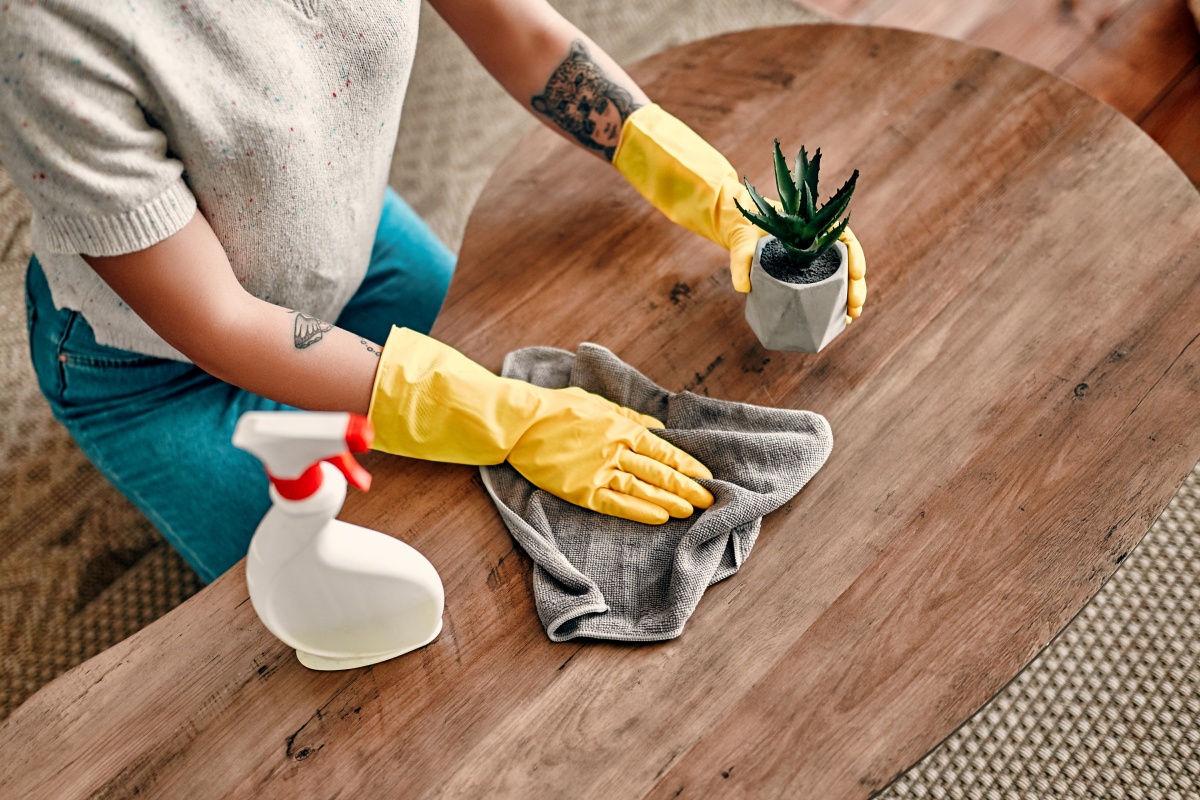 Person mit gelben Handschuhen hebt einen kleinen Blumentopf an, um den Holztisch mit einem Tuch zu reinigen.