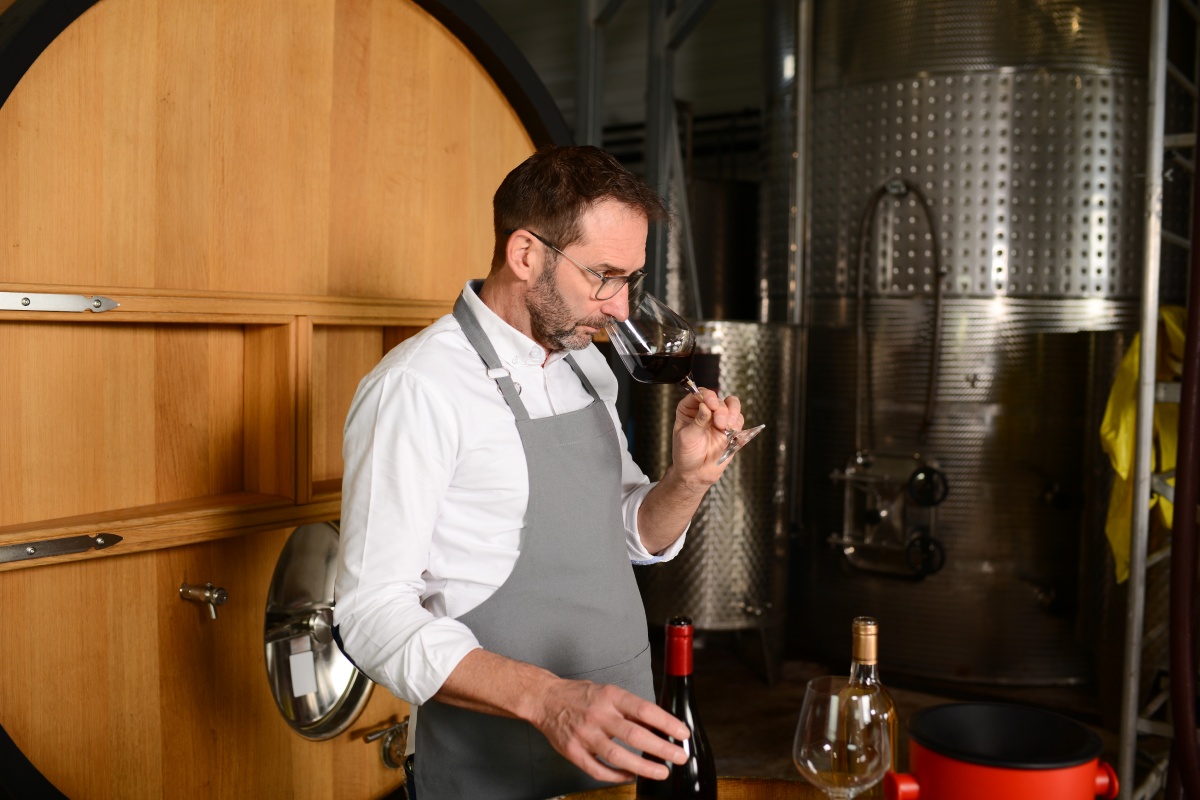 Ein Winzer in Schürze riecht in ein Glas Rotwein. Hinter ihm ein Holzfass und Edelstahlbehälter in einer Weinkellerei.