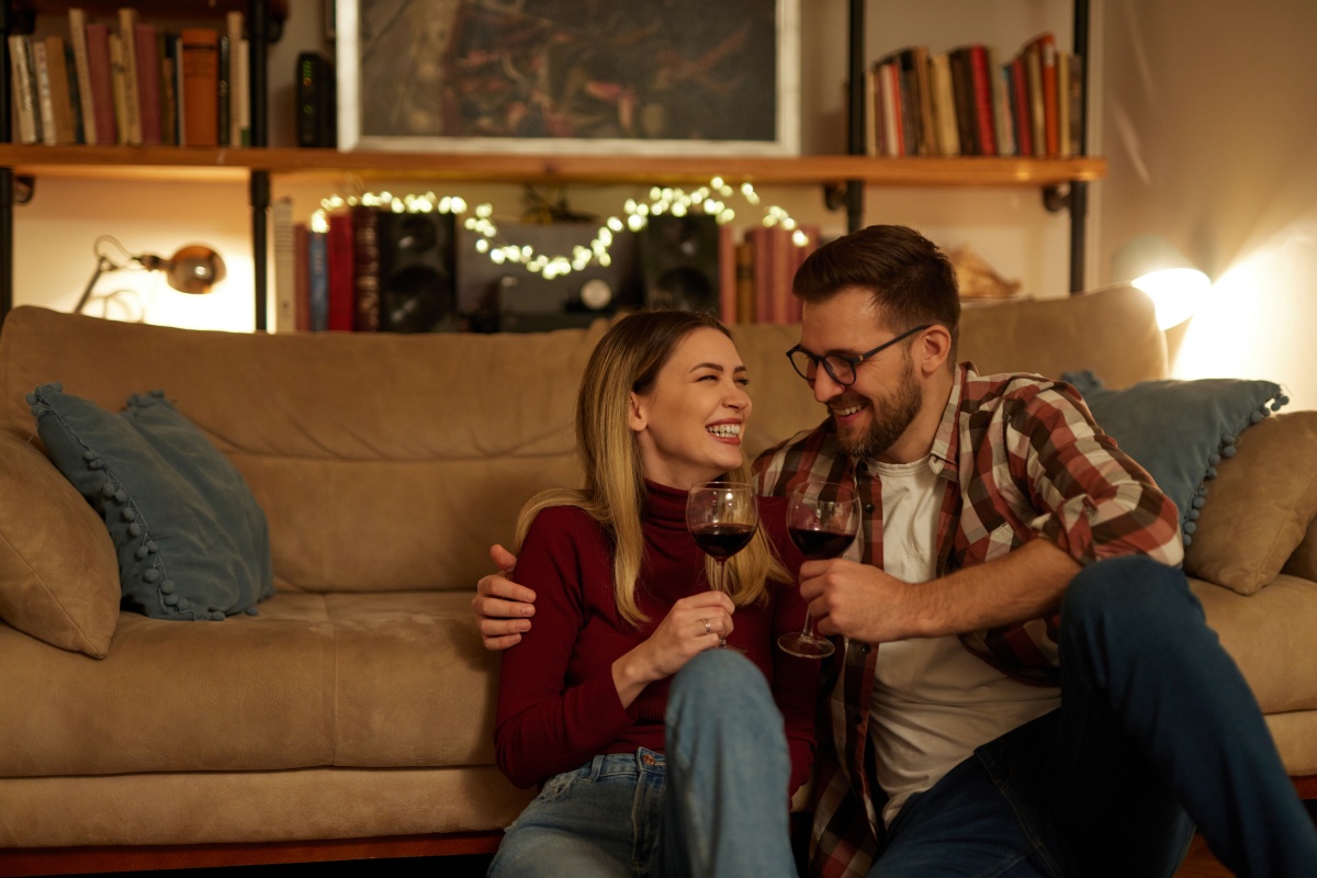 Ein Paar sitzt lachend auf dem Boden vor einem Sofa in einem gemütlichen Wohnzimmer mit gedimmtem Licht. Sie halten Rotweingläser in ihren Händen.