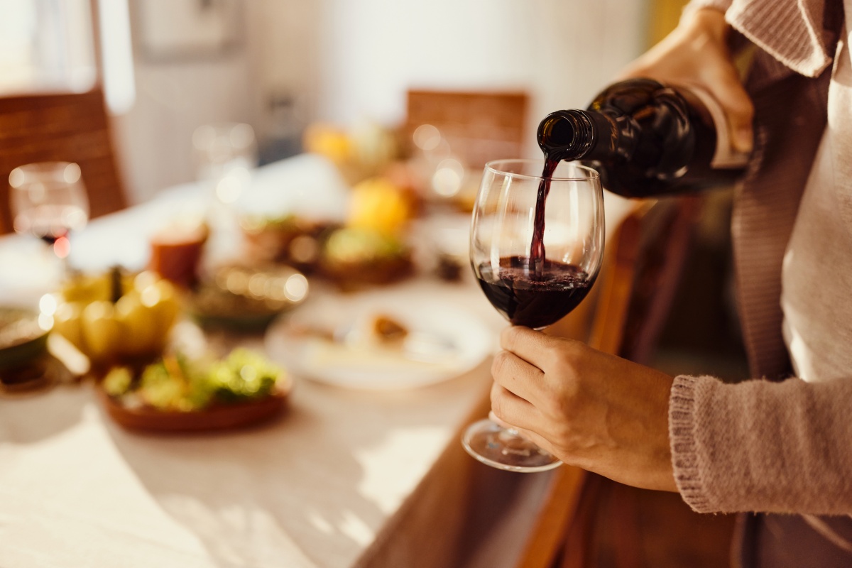Eine Person schenkt Rotwein in ein Glas ein, im Hintergrund ein gedeckter Esstisch mit verschiedenen Speisen.