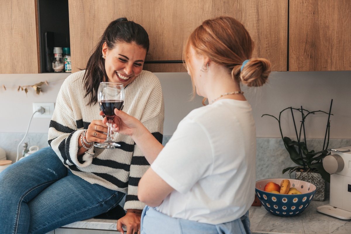 Zwei Frauen in einer Küche stoßen lächelnd mit Rotweingläsern an.