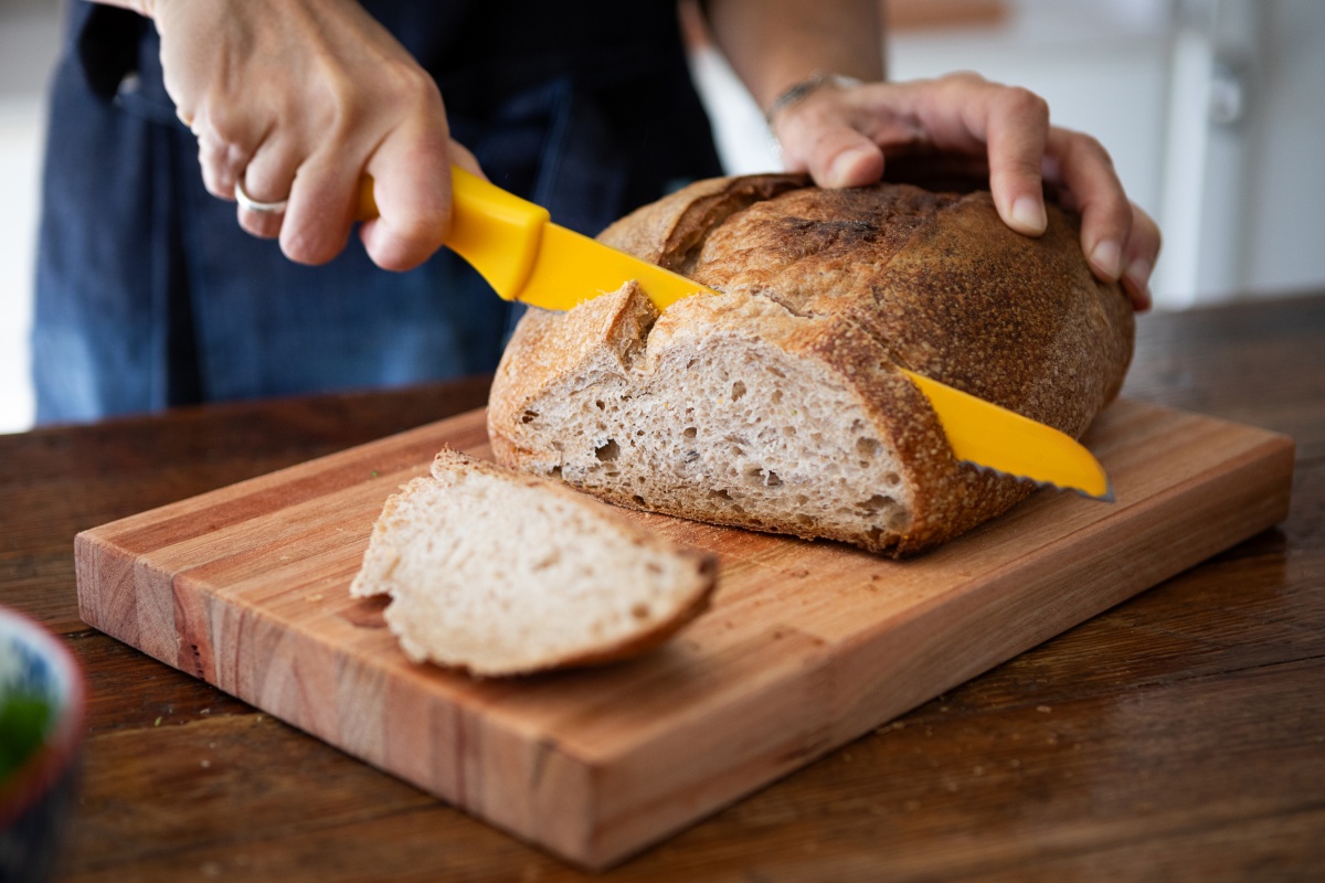 Eine Person schneidet mit einem gelben Brotmesser frisch gebackenes Sauerteigbrot auf einem Holzbrett.