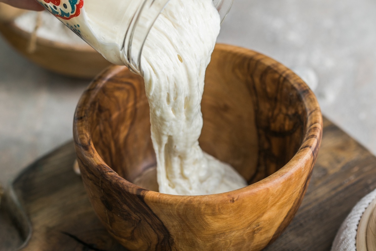 Person gießt Sauerteig von einem Glas in eine Holzschüssel.