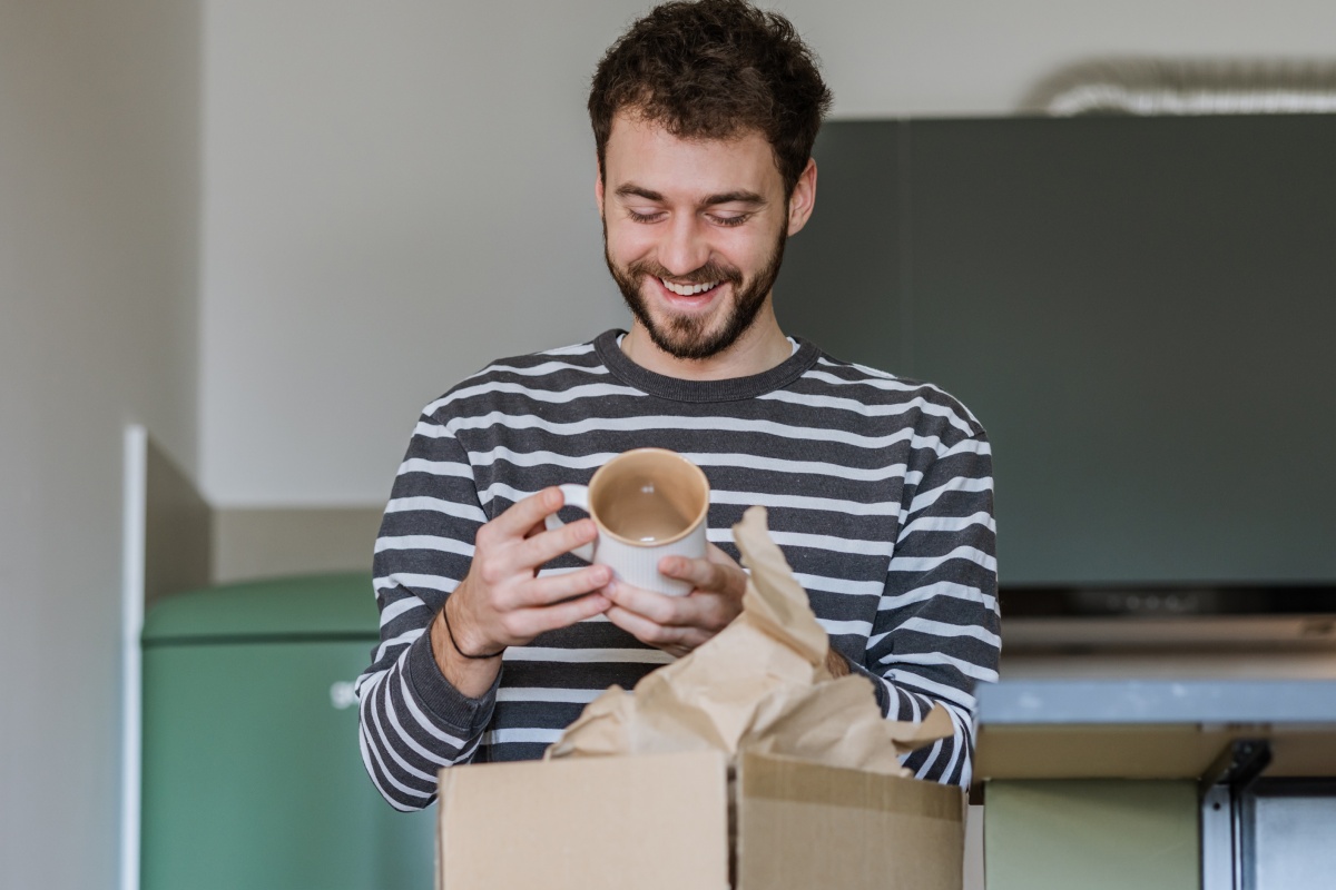 Mann packt eine Tasse aus einem Karton aus und lächelt.