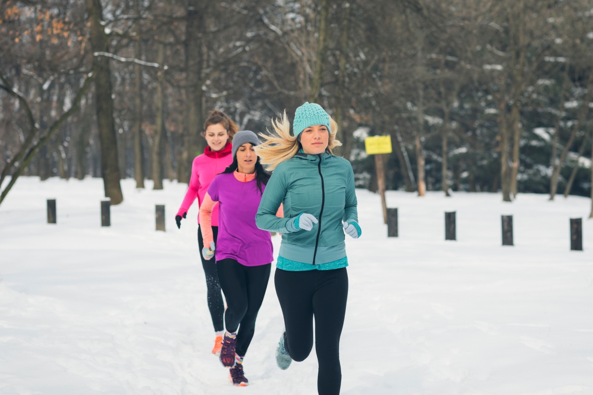 Drei Frauen joggen in bunten Jacken hintereinander durch einen verschneiten Park.