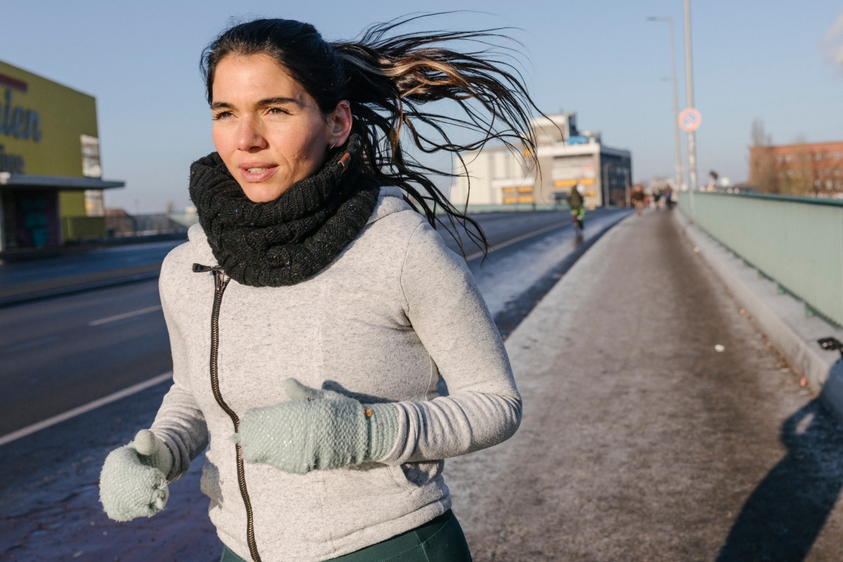 Frau joggt an einem sonnigen Wintertag über eine Brücke. Sie trägt Schal, Handschuhe und Sportkleidung.