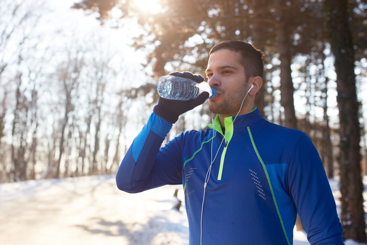 Mann mit blauer Laufjacke und Kopfhörern steht auf einem verschneiten Waldweg und trinkt Wasser aus einer Flasche.