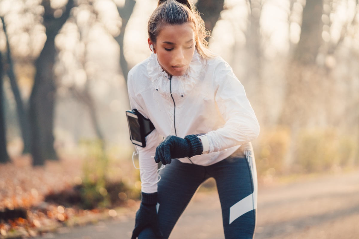 Frau in weißer Jacke überprüft ihre Smartwatch bei kurzer Pause während des Joggens im Park.