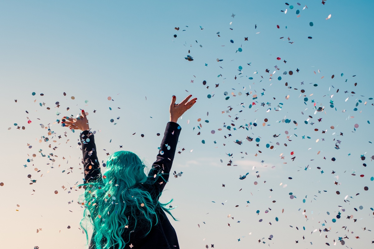 Person mit langen, türkisfarbenen Haaren wirft Konfetti in die Luft bei blauem Himmel im Freien.