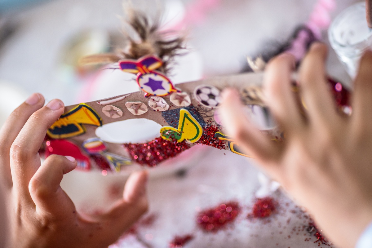 Person hält eine Maske mit Glitzer, Stickern und Federn in den Händen auf einem Basteltisch.
