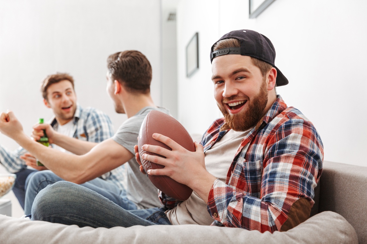 Lachender Mann mit American-Football in der Hand, sitzt auf einem Sofa, während Freunde im Hintergrund jubeln.