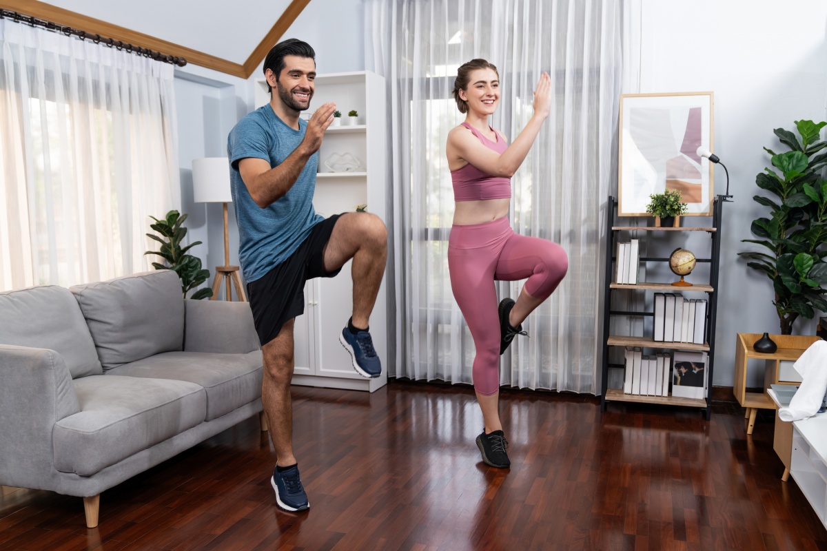 Mann und Frau trainieren gemeinsam mit Kniehebe-Übungen im Wohnzimmer zu Hause vor einem Sofa.