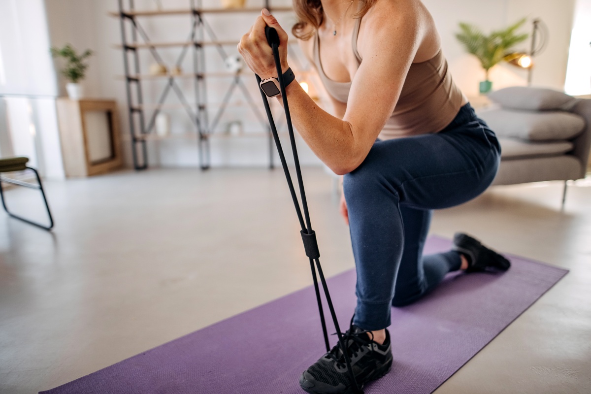 Frau kniet auf einer lila Yogamatte und trainiert mit einem Fitnessband in einem großen Wohnzimmer zu Hause.