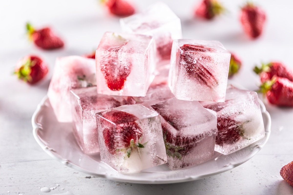 Eiswürfel mit eingefrorenen Erdbeeren auf einem weißen Teller, daneben frische Erdbeeren.