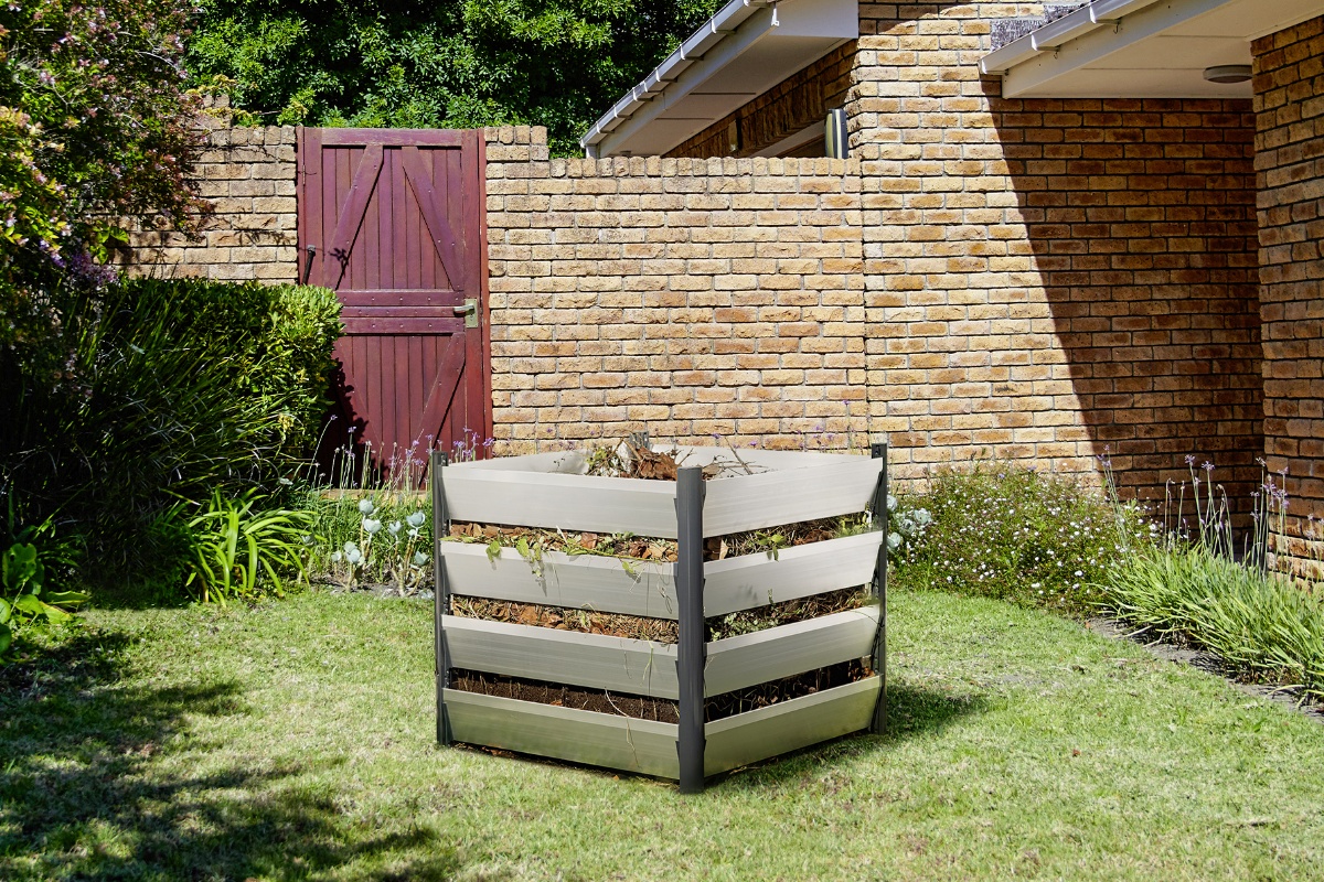 Metall-Komposter in einem Garten mit am Rand schmal angelegtem Blumenbeet, dahinter Hauswand aus Backsteinen mit Holztür.