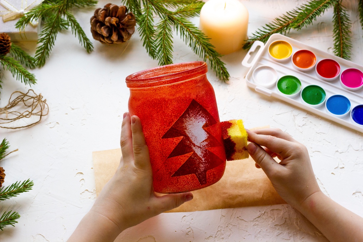 Eine Person betupft ein altes Vorratsglas mit roter Farbe und Tannenmotiv, dahinter ein Farbkasten, Tannenzweige und eine Kerze.
